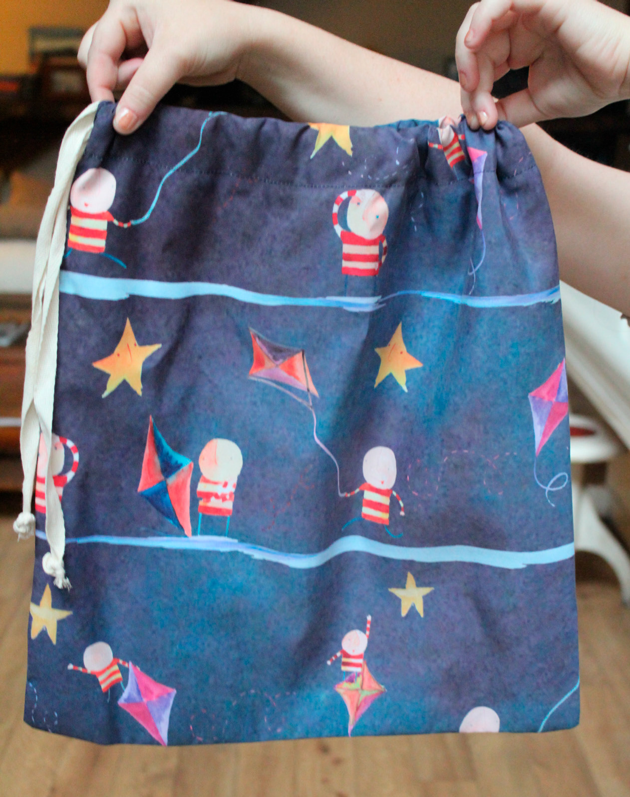 A child's hands holding up a navy drawstring bag printed with the Bellita pattern of kites, stars and striped explorers